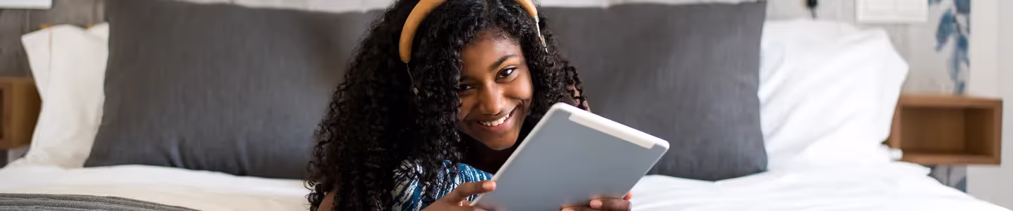 Uma garota feliz deita na cama, imersa em sua alegria, usando fones de ouvido enquanto explora animadamente seu tablet. Uma cena de descontração e contentamento.