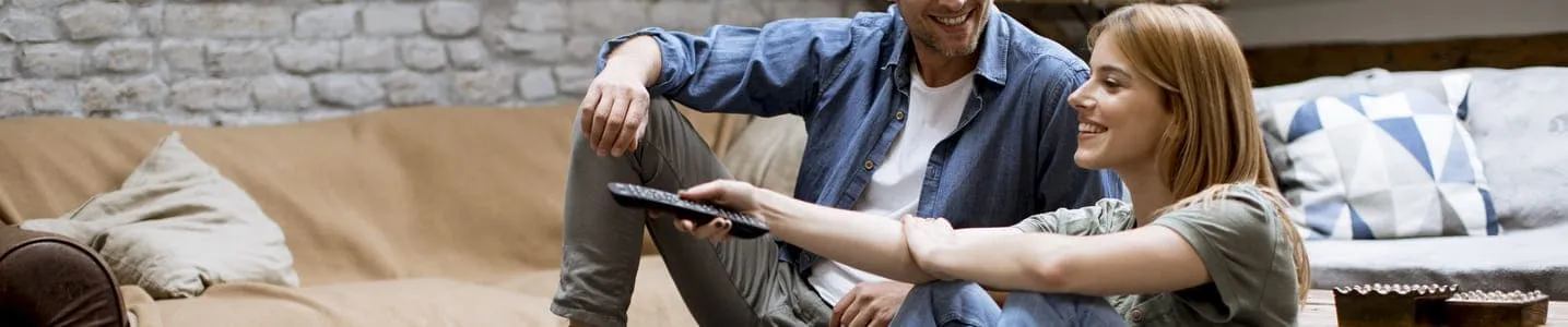 O casal, de forma descontraída, senta-se juntos no sofá, navegando pelas opções, tentando decidir o que vão assistir, compartilhando risadas e momentos de cumplicidade.