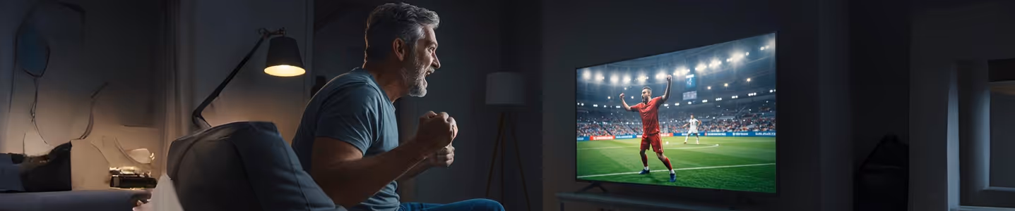 Homem vibrando assistindo jogo de futebol na TV em casa à noite.