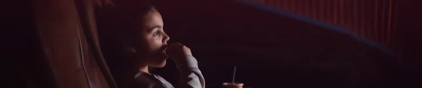 Criança saboreando pipoca enquanto se diverte assistindo algo com atenção.