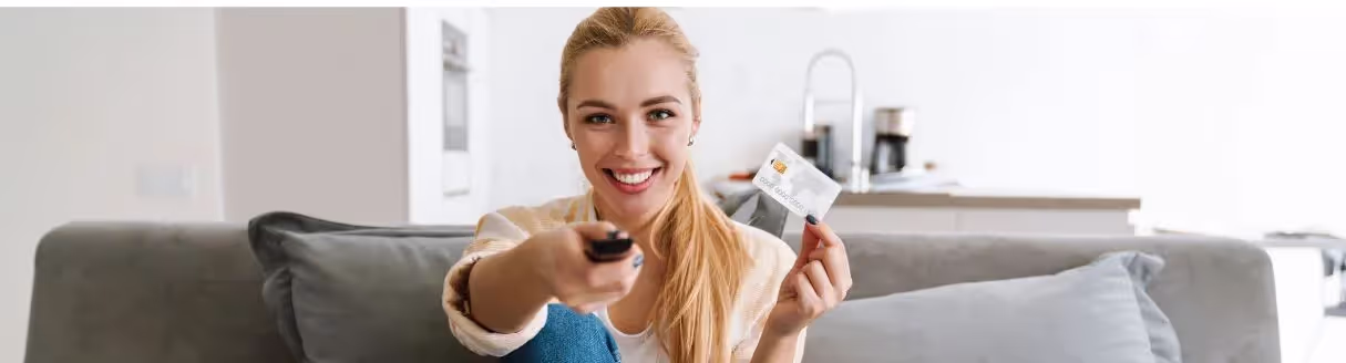 Com um sorriso no rosto, a mulher segura seu cartão de crédito enquanto assiste TV com o controle remoto, desfrutando de um momento de lazer e conforto em casa.