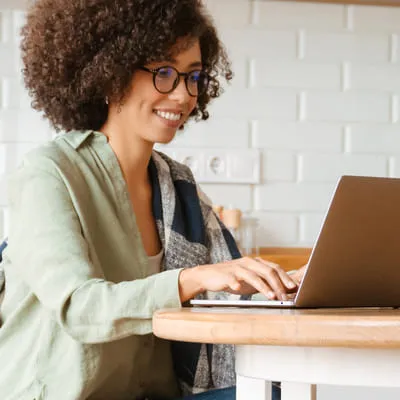 Com um sorriso, a jovem mulher desfruta de um momento de trabalho no notebook em sua cozinha, combinando eficiência e conforto.