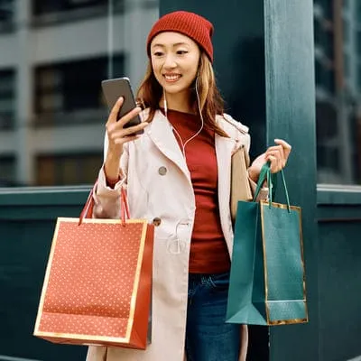 Com sacolas de compras em mãos, a jovem mulher expressa alegria ao conferir seu celular, talvez celebrando uma compra bem-sucedida ou compartilhando a novidade com amigos.