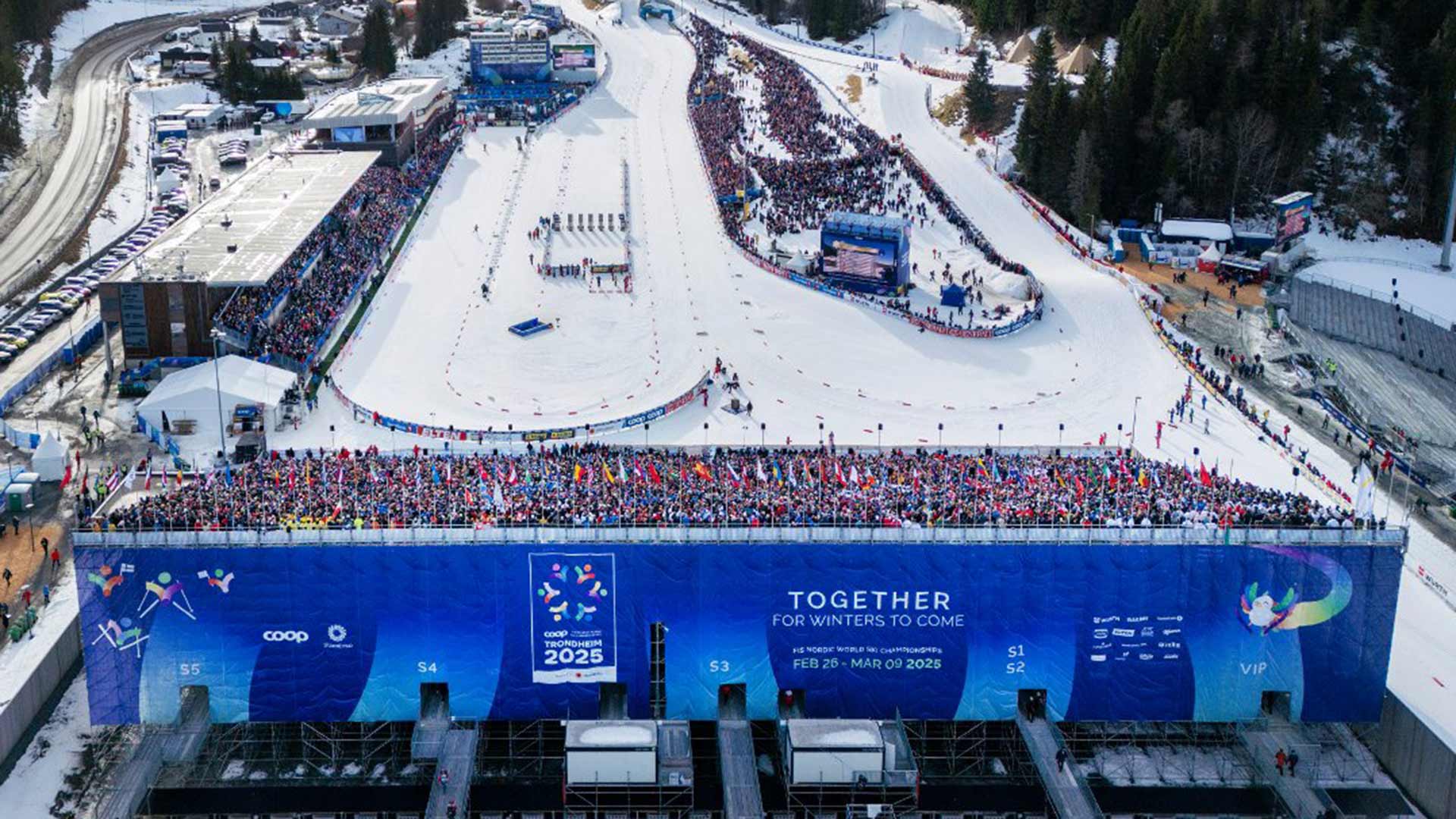 Luftfoto av et vintersportsarena fylt med et stort publikum, med skiløyper og arrangement for ski-VM i Trondheim 2025 annonsert på en blå banner.