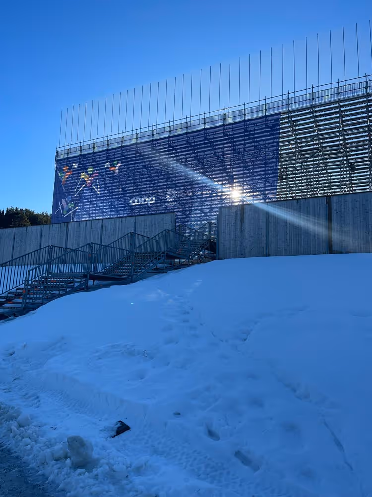 Tribunestruktur med banner som viser skiløpere, snø på bakken og sol som skinner mot tribunen.