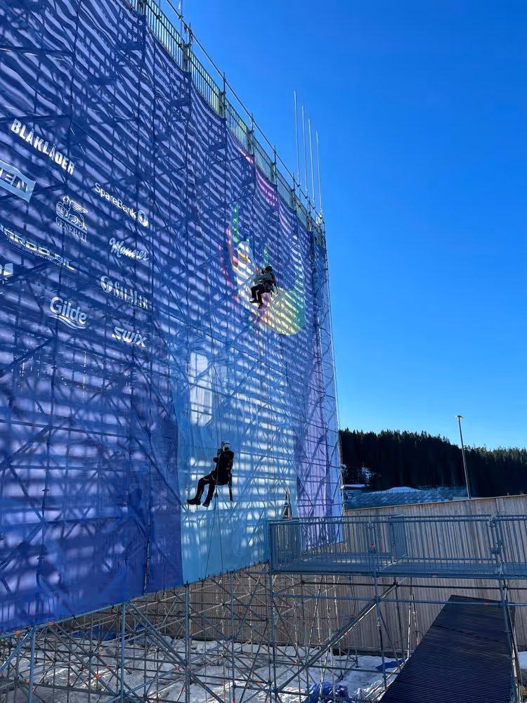 To arbeidere med sikkerhetsutstyr klatrer på stillaset til et høyt banner under en klar blå himmel.