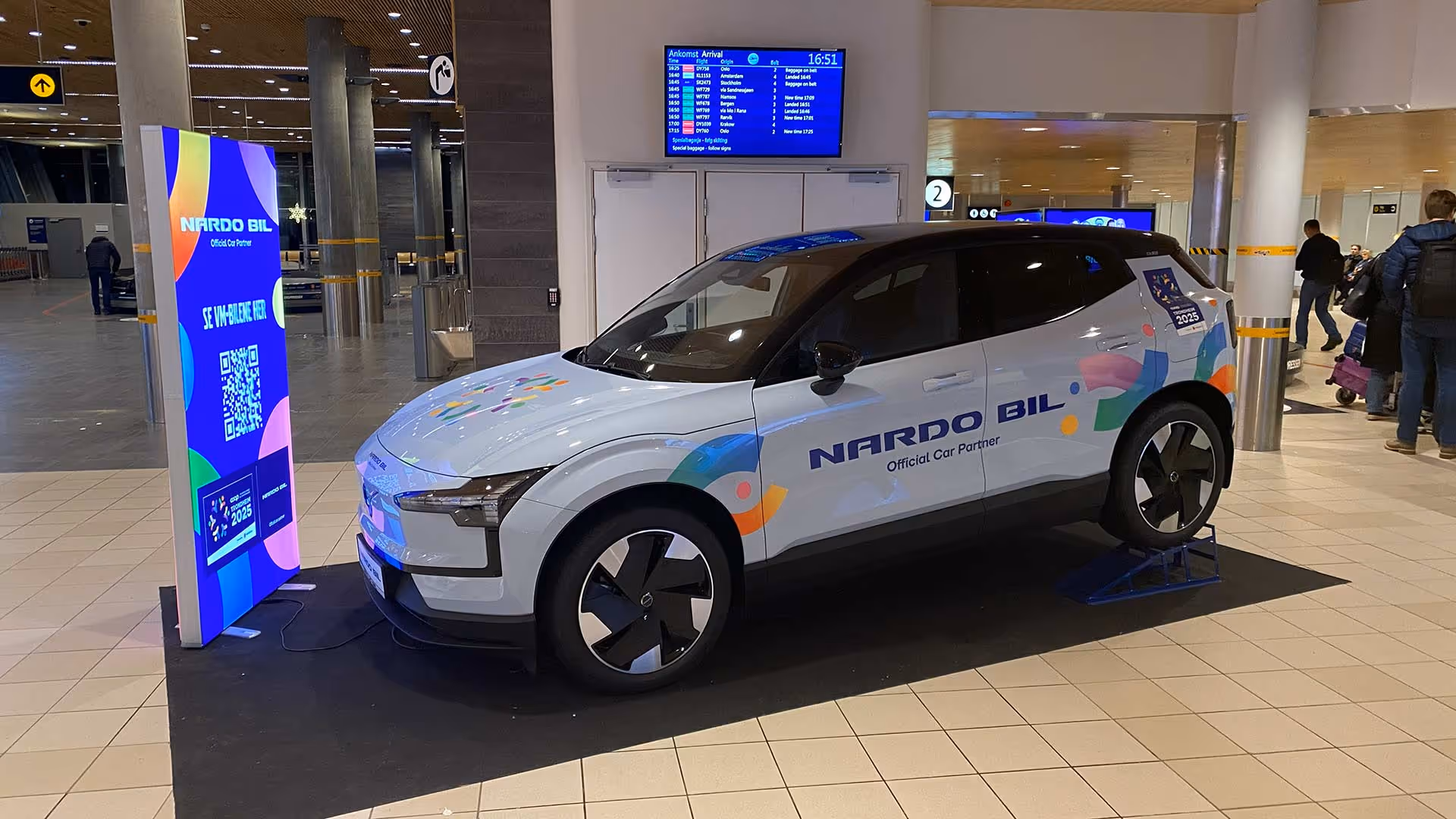 Hvit Polestar elbil med NARDO BIL-logo og fargerike dekorasjoner utstilt innendørs på et flyplassområde.