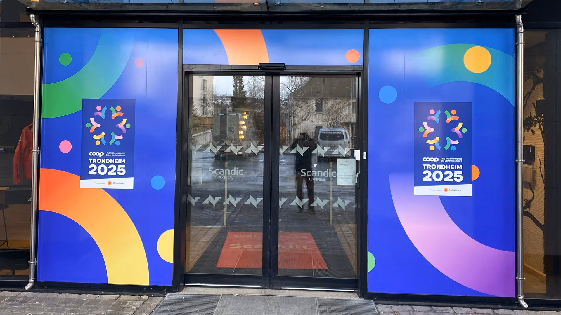 Glass entrance doors of Scandic Lerkendal hotel with colorful posters promoting Coop FIS Nordic World Ski Championships Trondheim 2025.