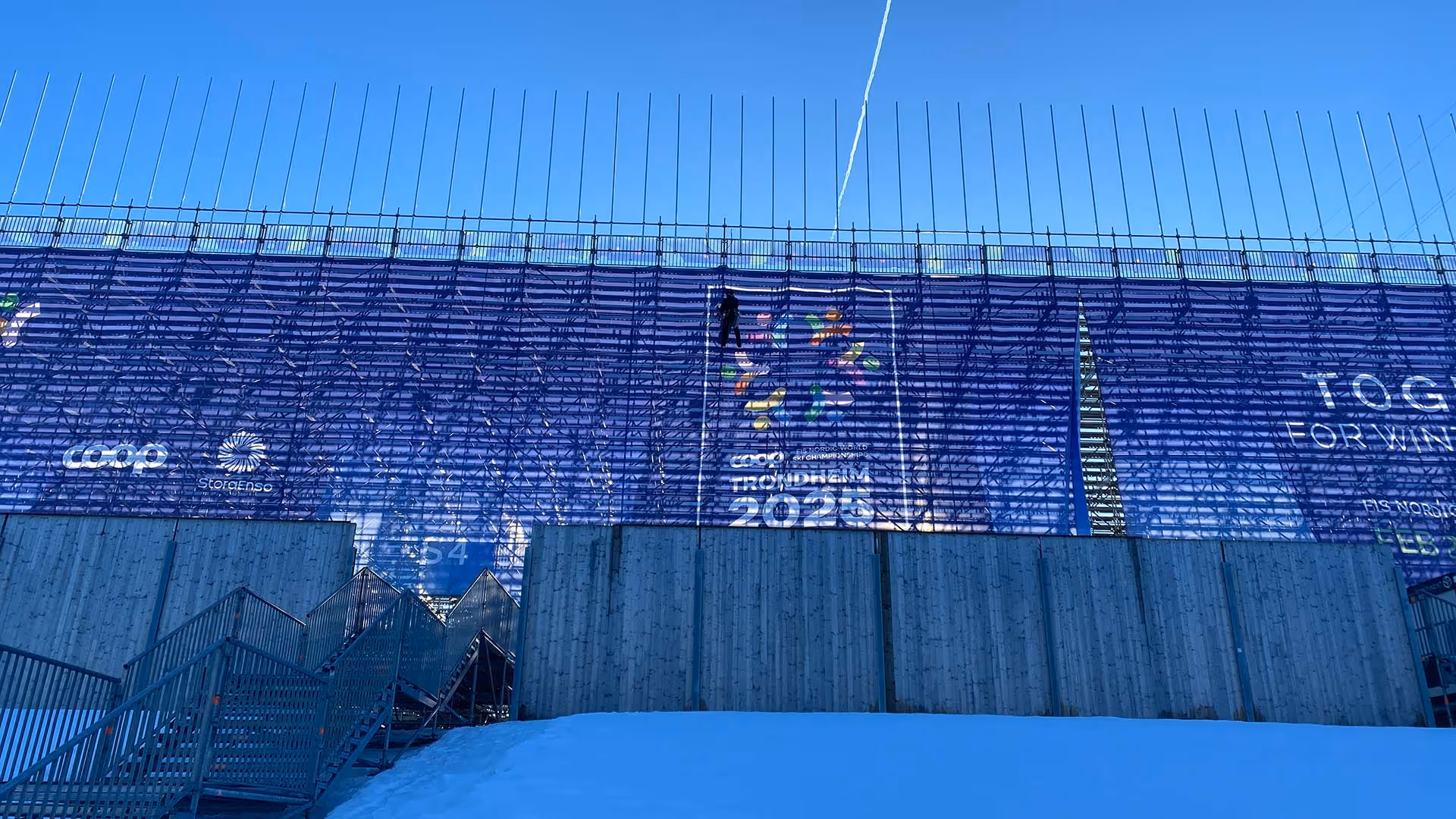 Person som arbeider i høyden på stillas foran et stort blått banner med logoer og teksten Trondheim 2025 under klar blå himmel.