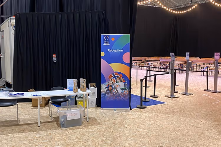 Mottak med bord og stoler foran et fargerikt skilt med teksten "Reception" og bilder av glade mennesker, i en stor hall med benker og black curtains i bakgrunnen.