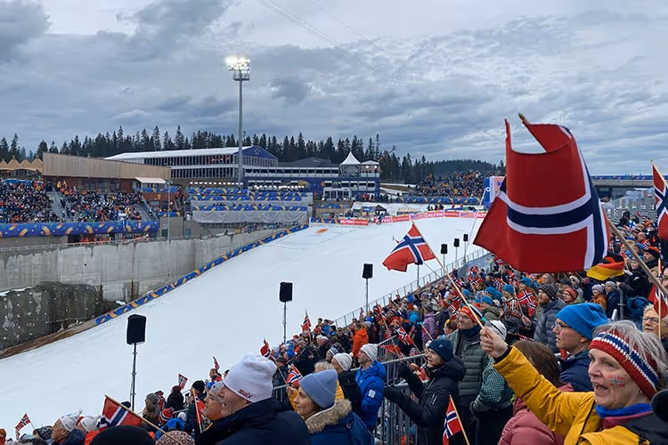 Tilstedeværelse av entusiastiske tilskuere som holder norske flagg og heier under et vinteridrettsarrangement ved en snødekt stadion.