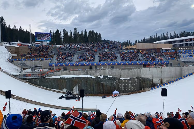 Folkemengde med norske flagg ser på et skiarrangement i en snødekt arena med tilskuere på tribunen og skog i bakgrunnen.