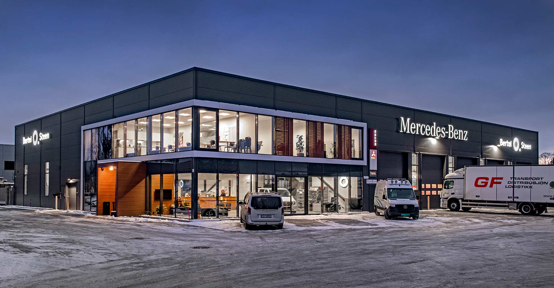 Moderne Mercedes-Benz-forhandler med kontor- og verkstedområde opplyst i skumringen og biler parkert utenfor på snødekt underlag.
