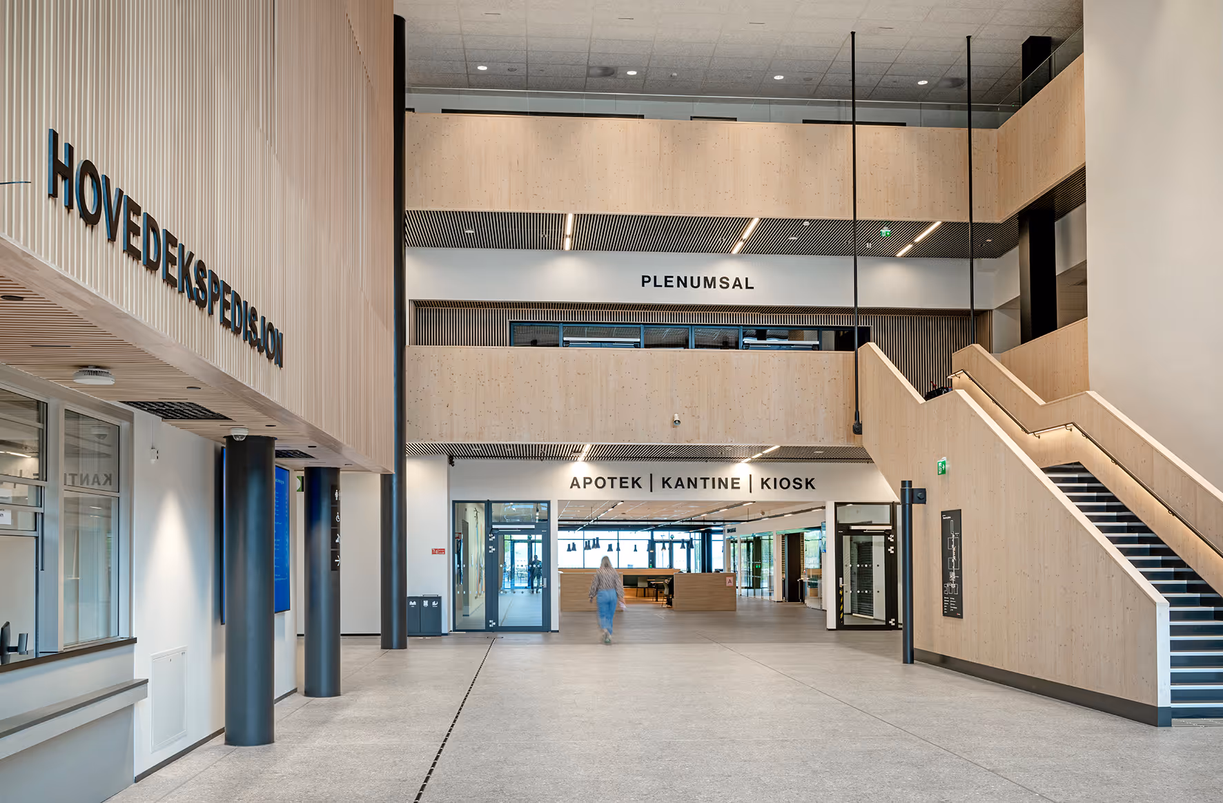Moderne sykehusentré med skilt for hovedekspedisjon, plenumsal, apotek, kantine og kiosk, samt en trapp til høyre.