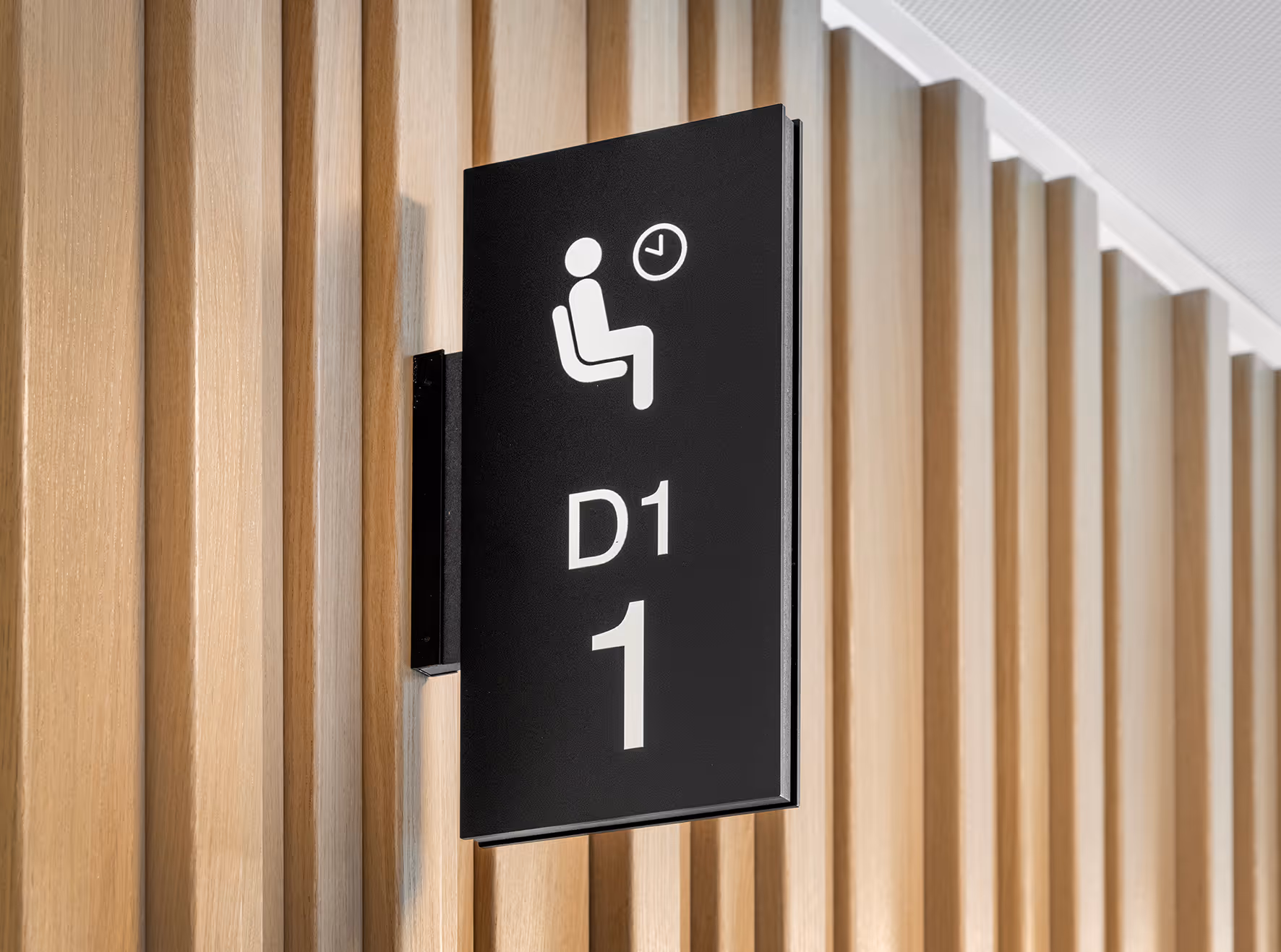 Skilt med et symbol av en sittende person og et klokkeikon over teksten D1 1, montert på en vegg med trepaneler.