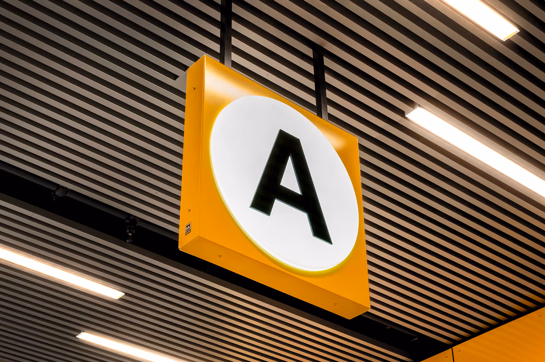 Gul og hvit skiltdel med bokstaven A hengende fra et mørkt tak med lysrør.