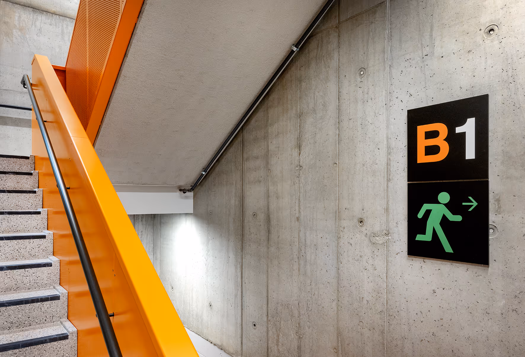 Trapp med oransje rekkverk ved siden av en betongvegg med skilt som viser B1 og nødutgangs-symbolet mot høyre.