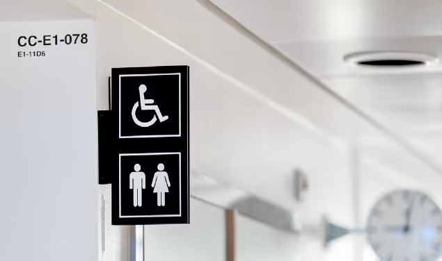 Skilt med symboler for rullestoltilgjengelighet og toalett for menn og kvinner på en vegg i en offentlig bygning.