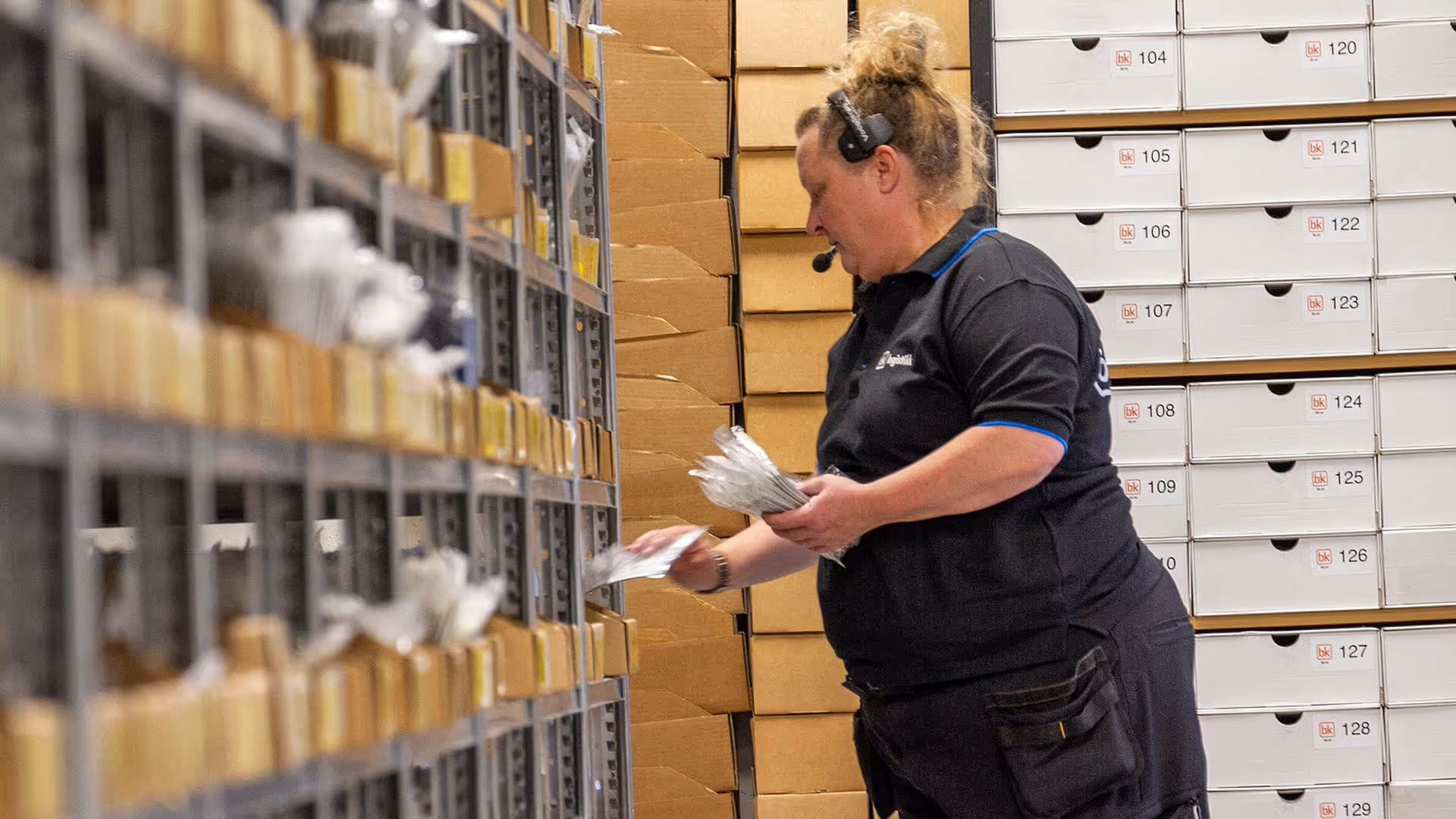 En kvinne med headset pakker små deler fra hyller i et lager med stablede esker.