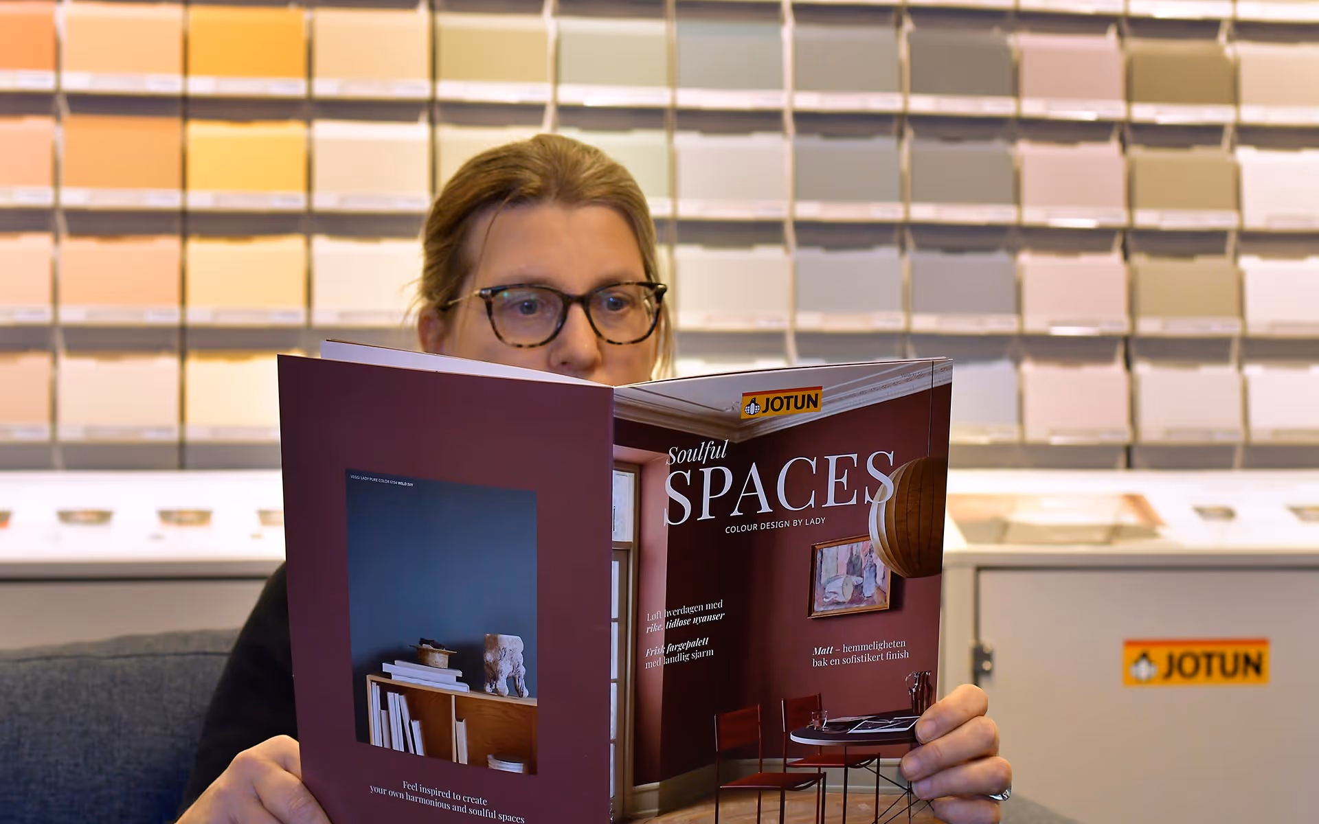 Person leser en Jotun fargebrosjyre foran en vegg med mange malingsprøver i ulike lyse nyanser.