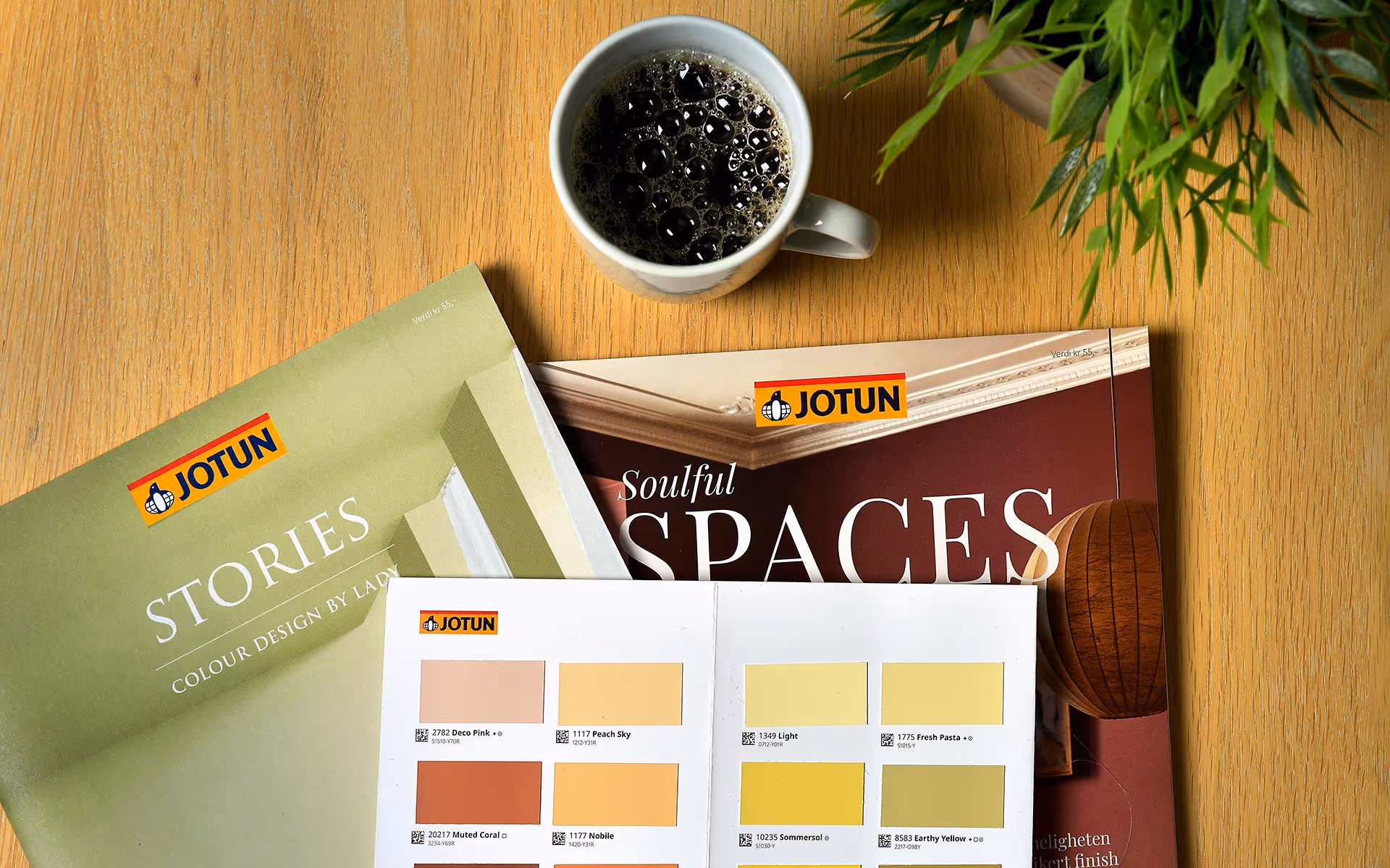 Oversikt over Jotun fargekart og magasiner på et trebord med en kopp svart kaffe og grønne planter øverst til høyre.