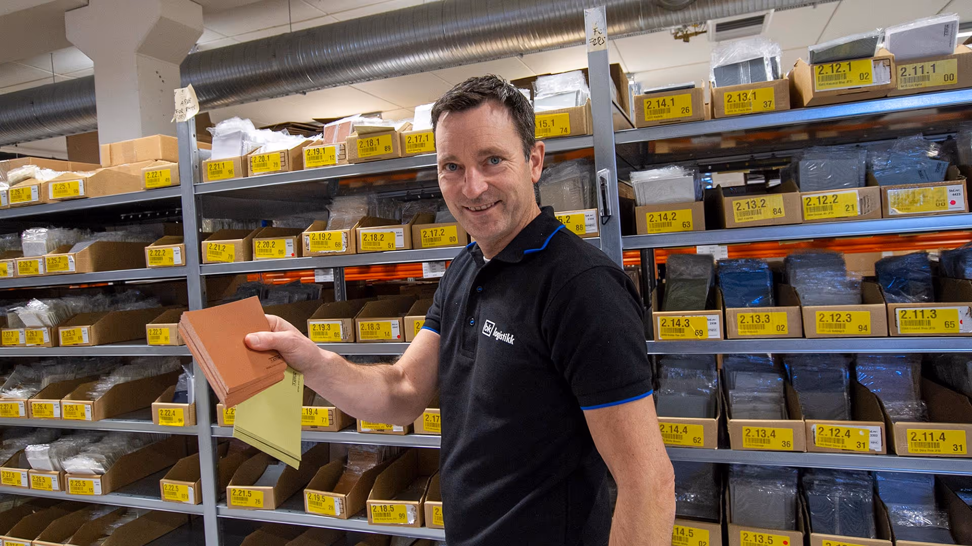 Mann i svart skjorte holder flere fargeprøver foran hyller med gule merkede esker i et lager.