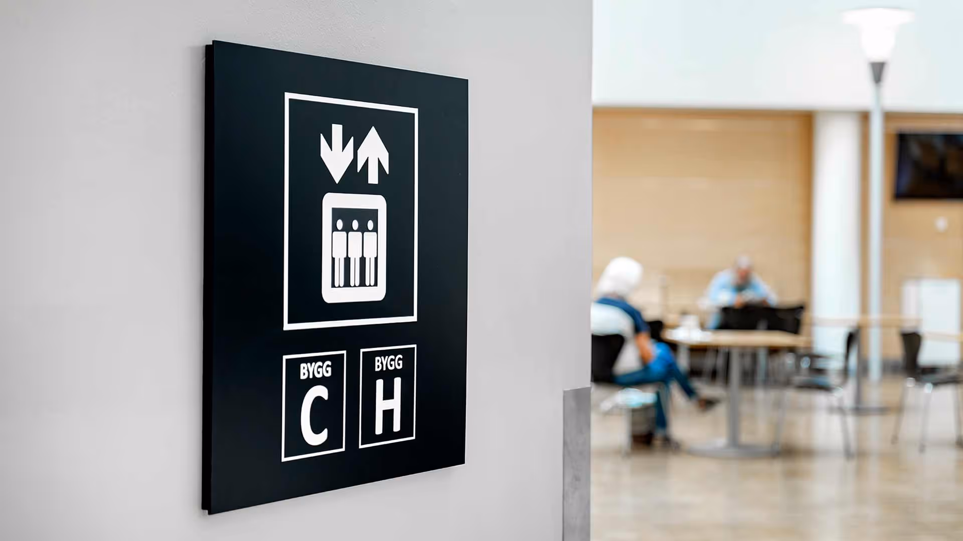 Skilt som viser heis med piler opp og ned, merket for Bygg C og Bygg H i en bygning.