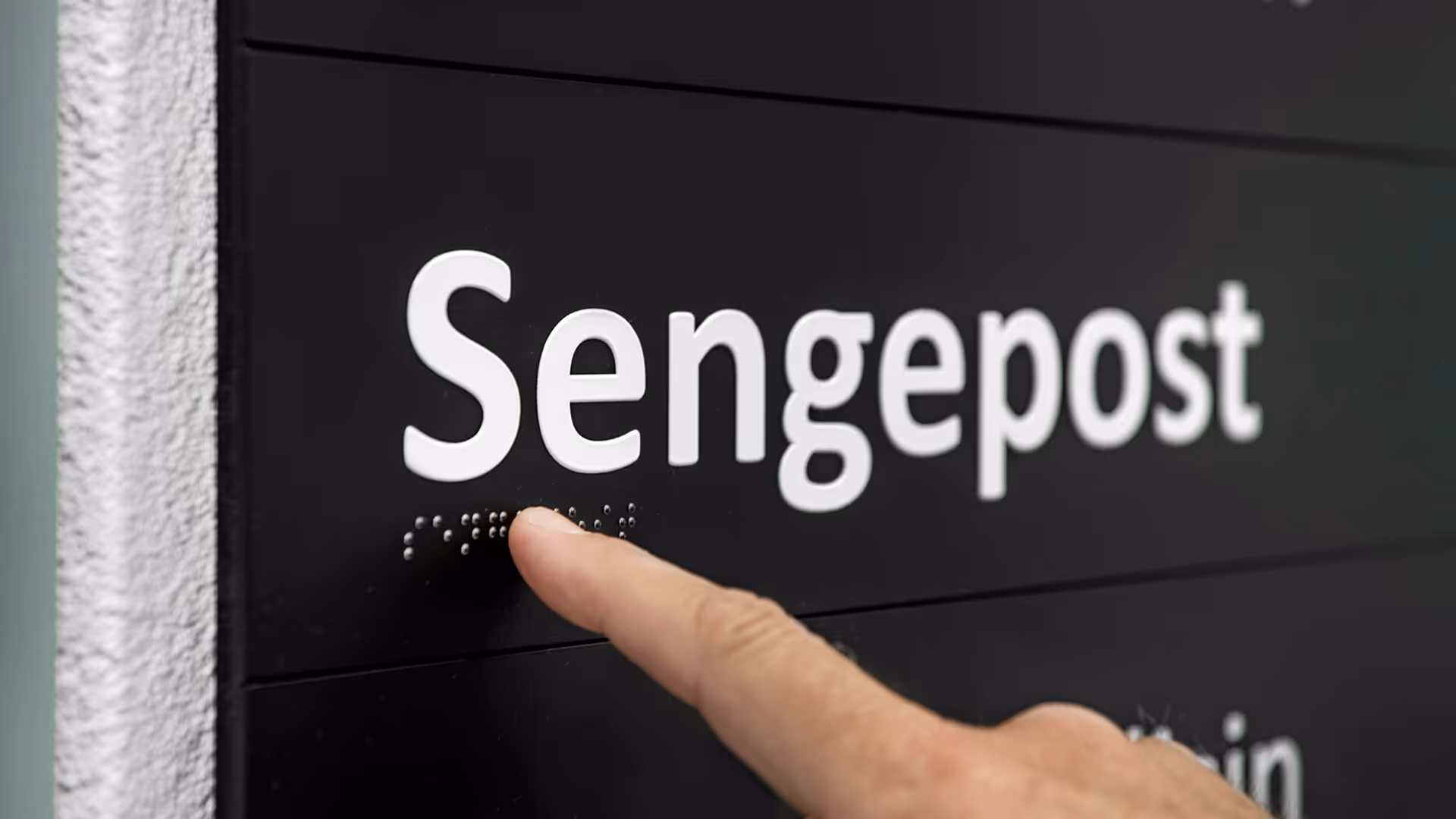 Fingertupp berører punktskrift under hvit tekst 'Sengepost' på svart skilt.