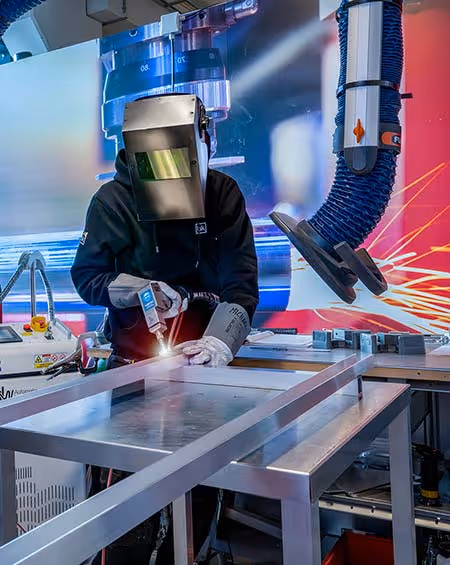 Person wearing welding helmet and gloves performing welding on metal beams in an industrial workshop.