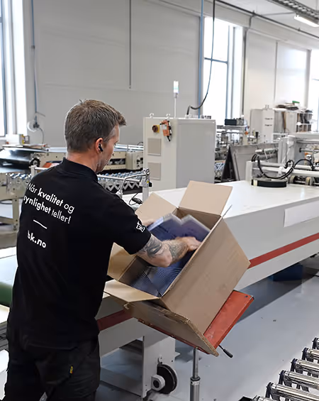 Mann pakker produkter i pappeske på fabrikkgulv med produksjonsmaskiner i bakgrunnen.