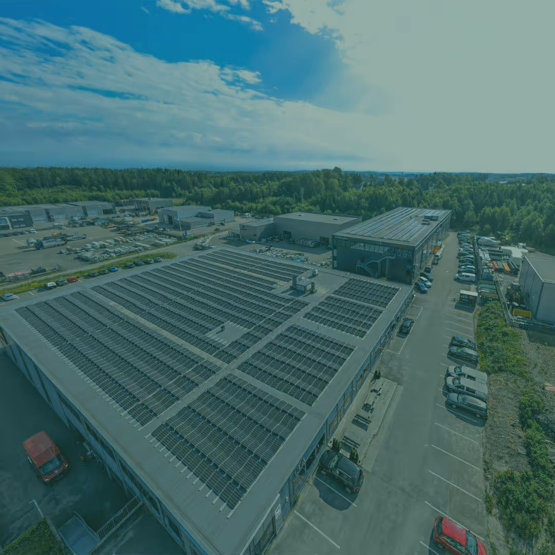 Luftfoto av et industriområde med flere bygninger, hvor taket på den nærmeste er dekket med solcellepaneler, omgitt av parkerte biler og grønt skogsområde i bakgrunnen.