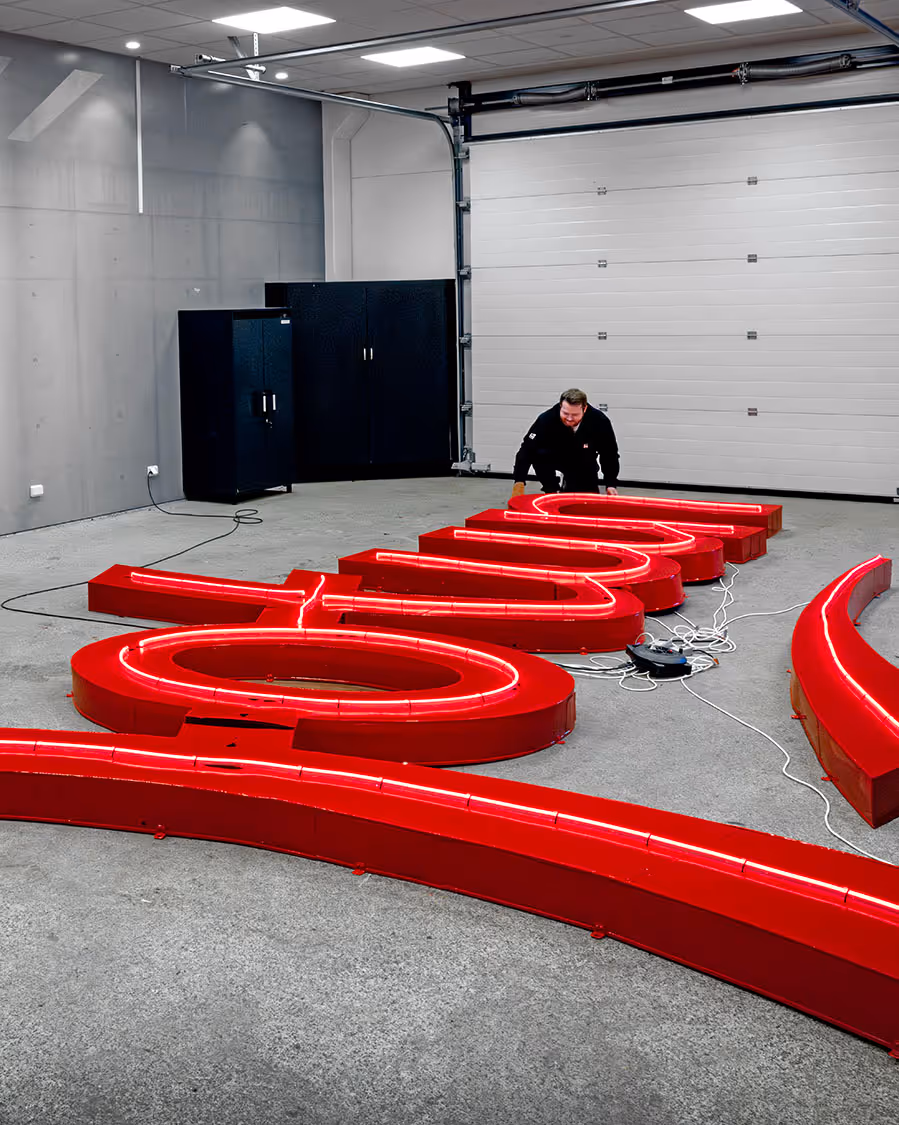 En person installerer store, røde opplyste bokstaver på gulvet i en grå garasje med lukket port.