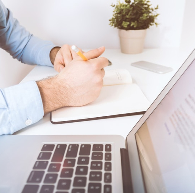 Persona con camicia azzurra che scrive su un taccuino con una matita, con un computer portatile e una pianta sullo sfondo.