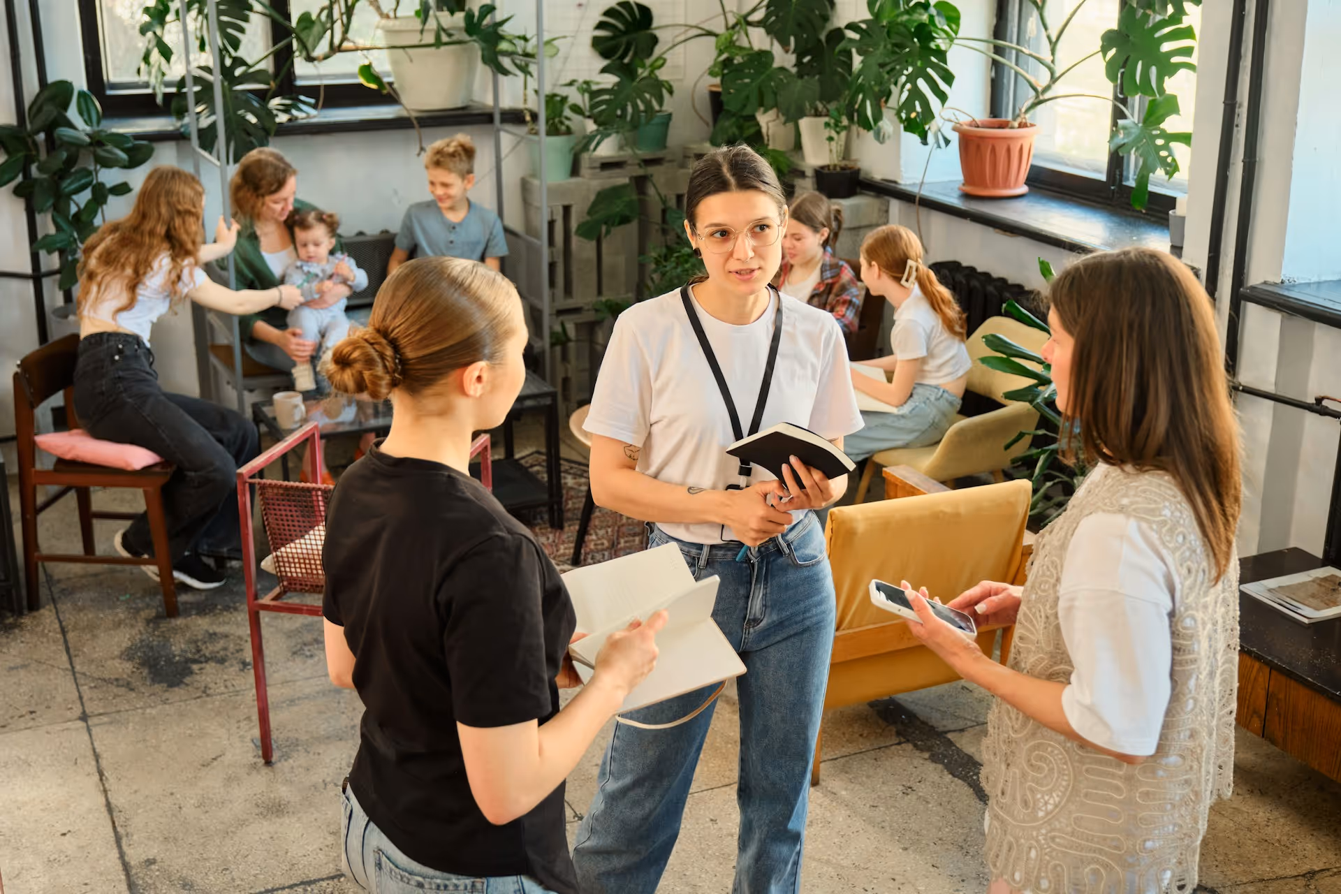 Un gruppo di persone in una stanza luminosa con molte piante, alcune conversano tenendo quaderni e uno smartphone, altre sono sedute interagendo.