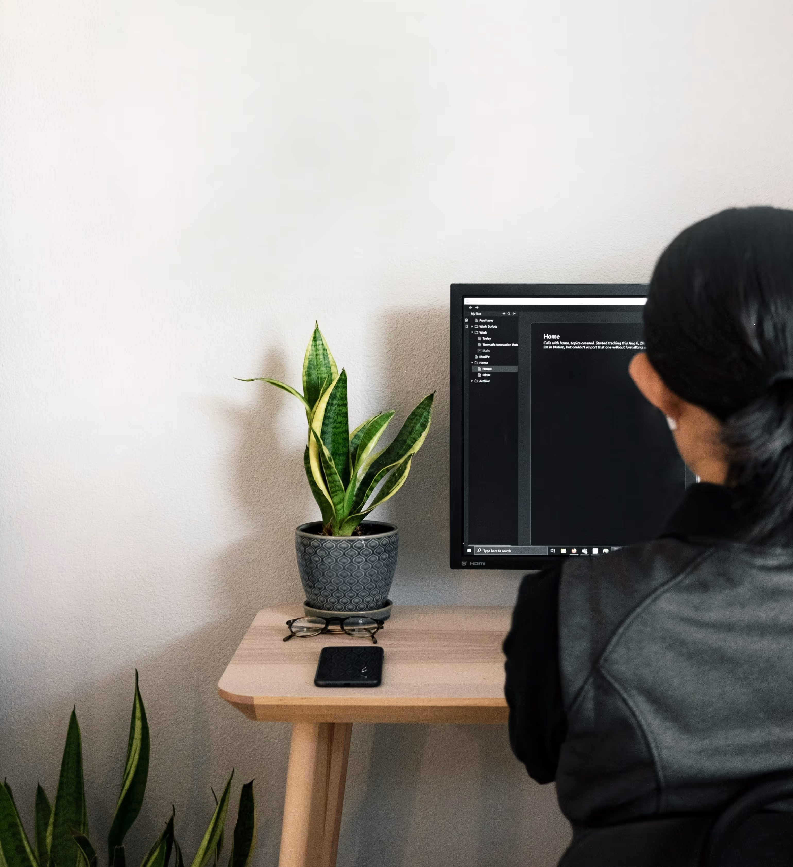 Persona con capelli legati che lavora al computer su una scrivania con pianta in vaso, occhiali e telefono.