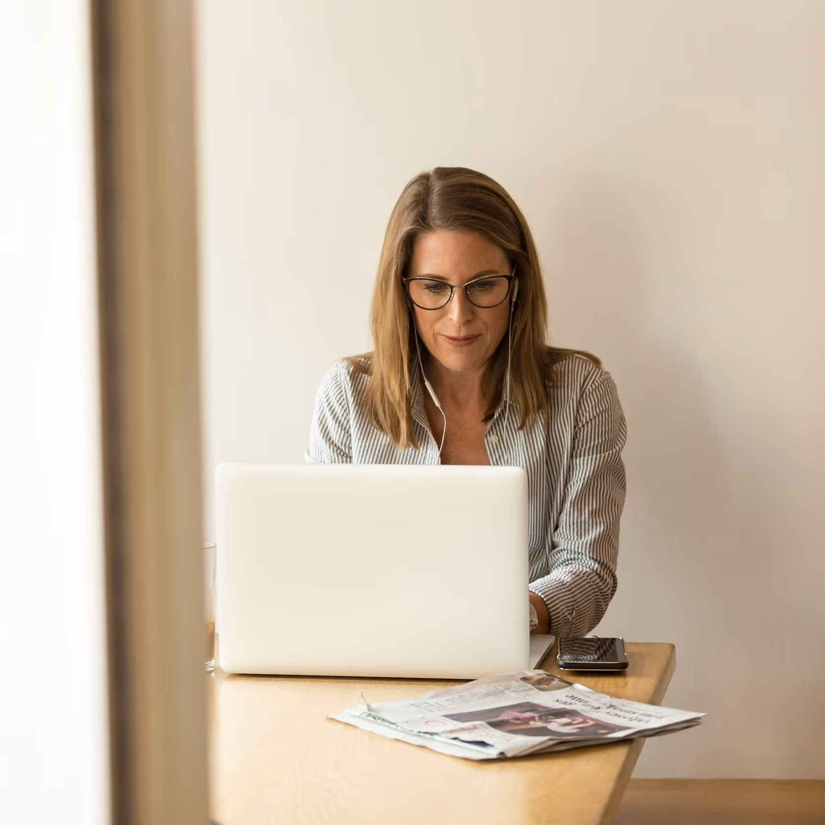 Donna con occhiali che usa un computer portatile mentre indossa auricolari bianchi, seduta a un tavolo con un giornale e uno smartphone.