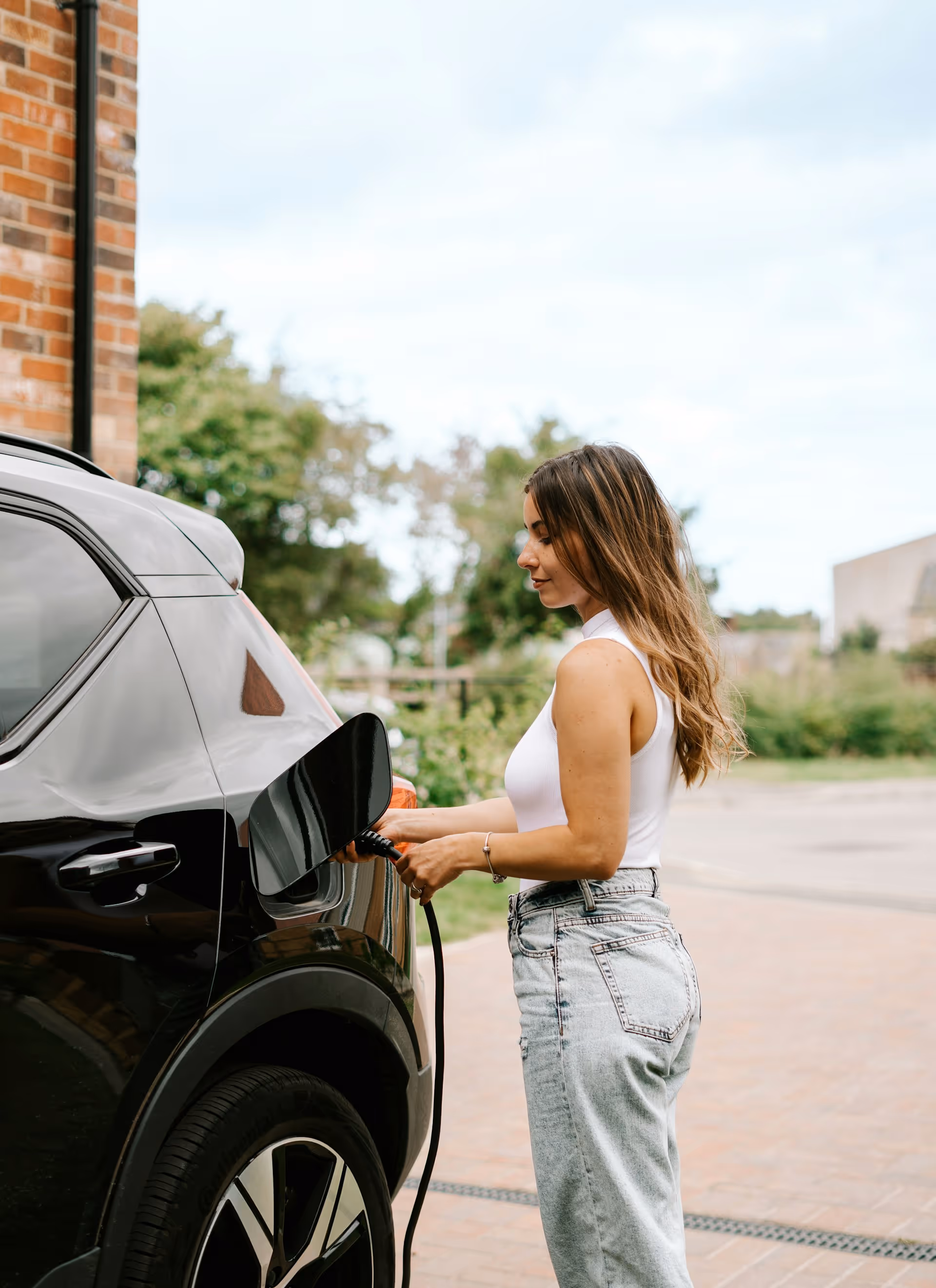 Donna che ricarica un'auto elettrica nera all'aperto vicino a un muro di mattoni.