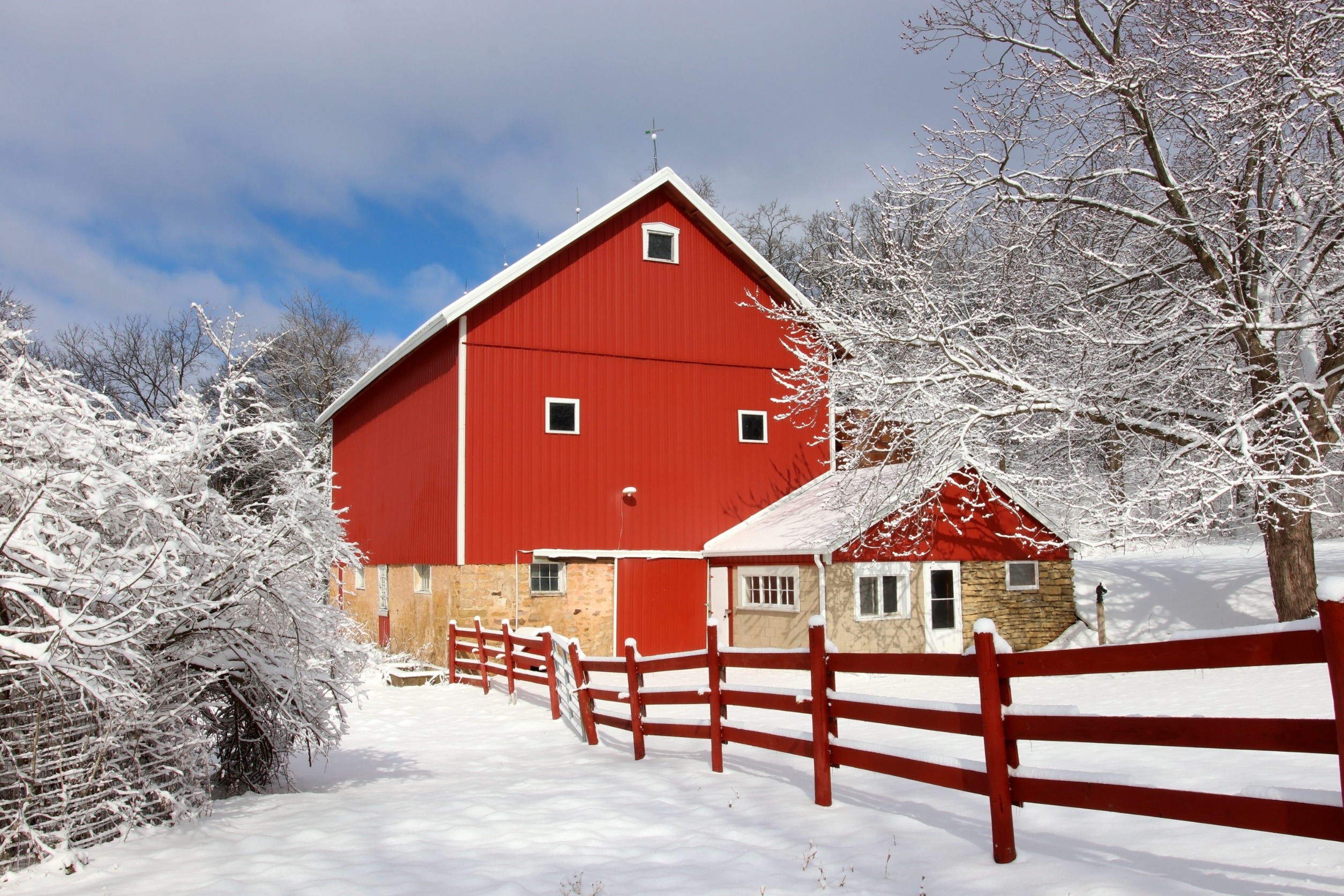 Red barn with stone base surrounded by snow-covered trees and a red fence under a partly cloudy sky.