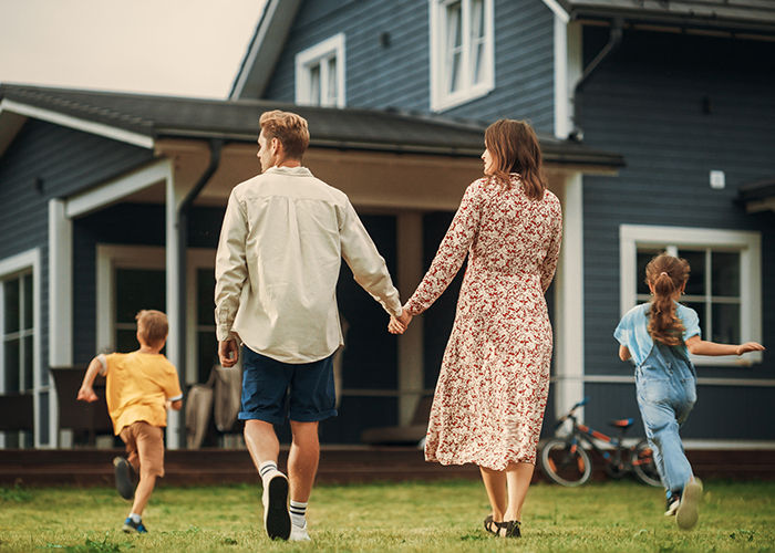 Couple holding hands walking on grass with two children running ahead near a blue house.