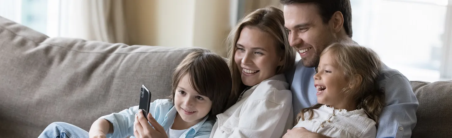 Smiling family of four sitting on a couch looking at a smartphone together.