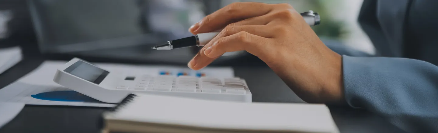 Person in a blue shirt using a white calculator with a pen in hand, with papers and a notebook on the table.