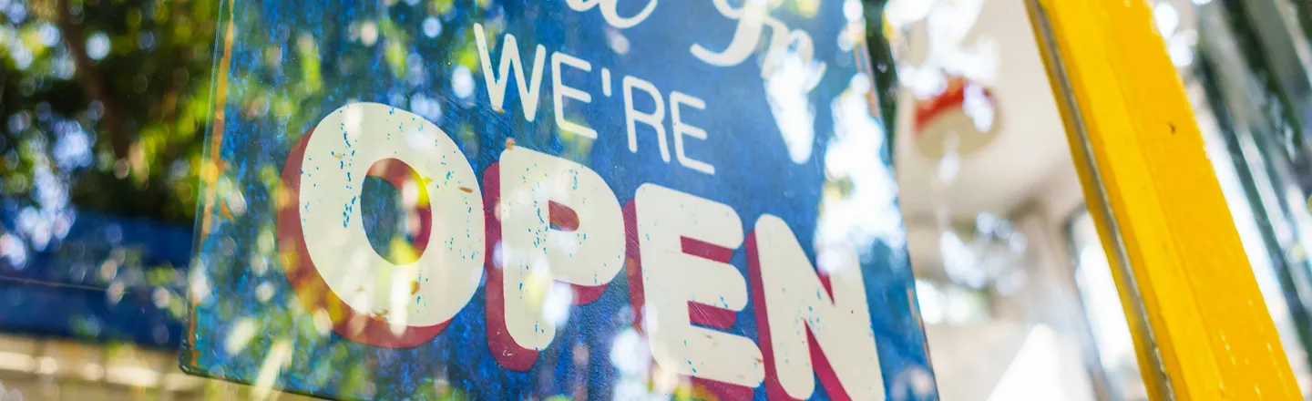 Blue sign on glass door with the text "WE'RE OPEN" in white letters, with reflections of trees and surroundings.