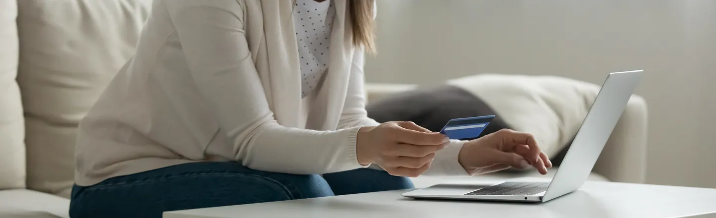 Person sitting on a couch holding a credit card while using a laptop on a white table.