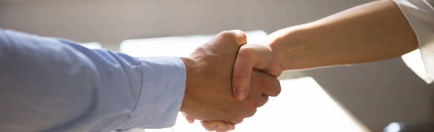 Close-up of two people shaking hands, one wearing a light blue shirt and the other a white sleeve, in a bright setting.