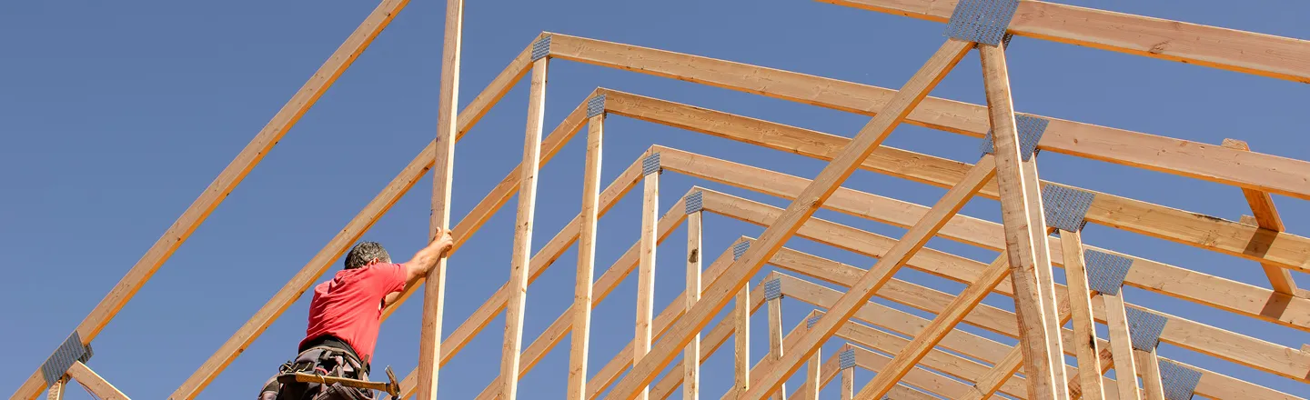 Construction worker in red shirt assembling wooden roof framework under clear blue sky.