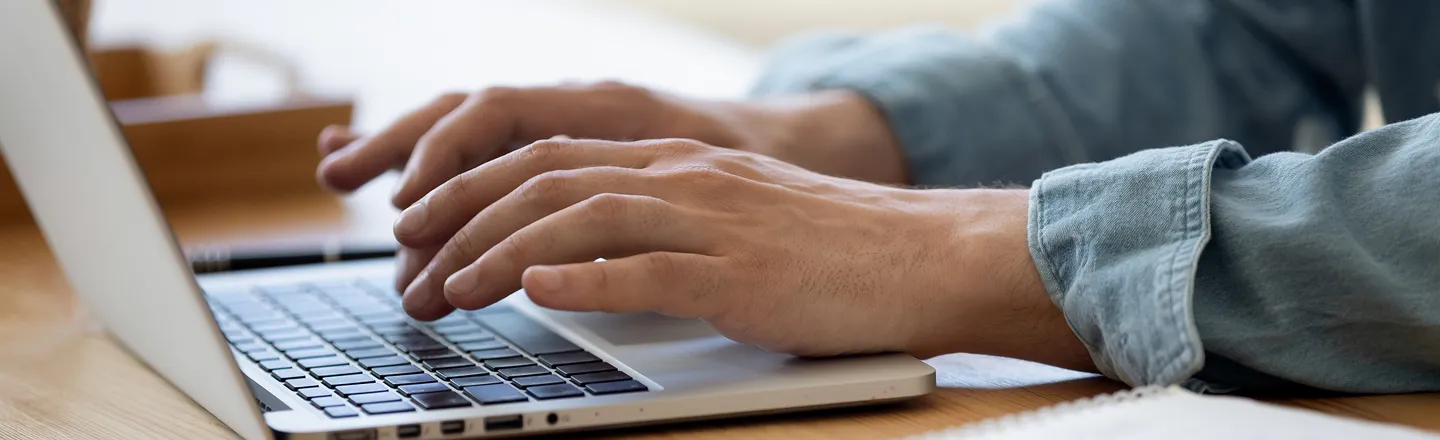 Close-up of hands typing on a laptop keyboard on a wooden desk with a notebook nearby.