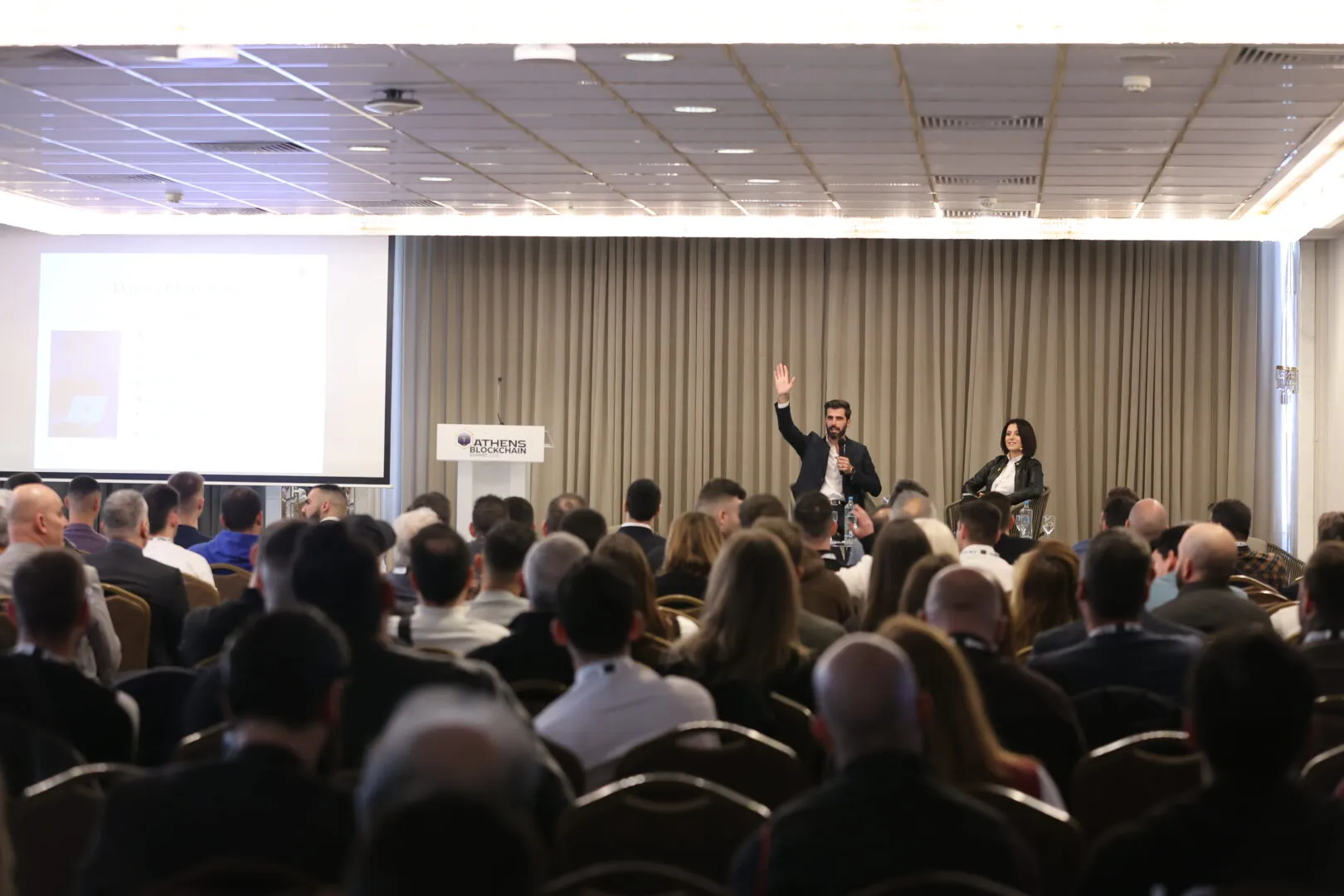 A man speaking and raising his hand on stage during a conference with a seated woman beside him and a large audience watching.