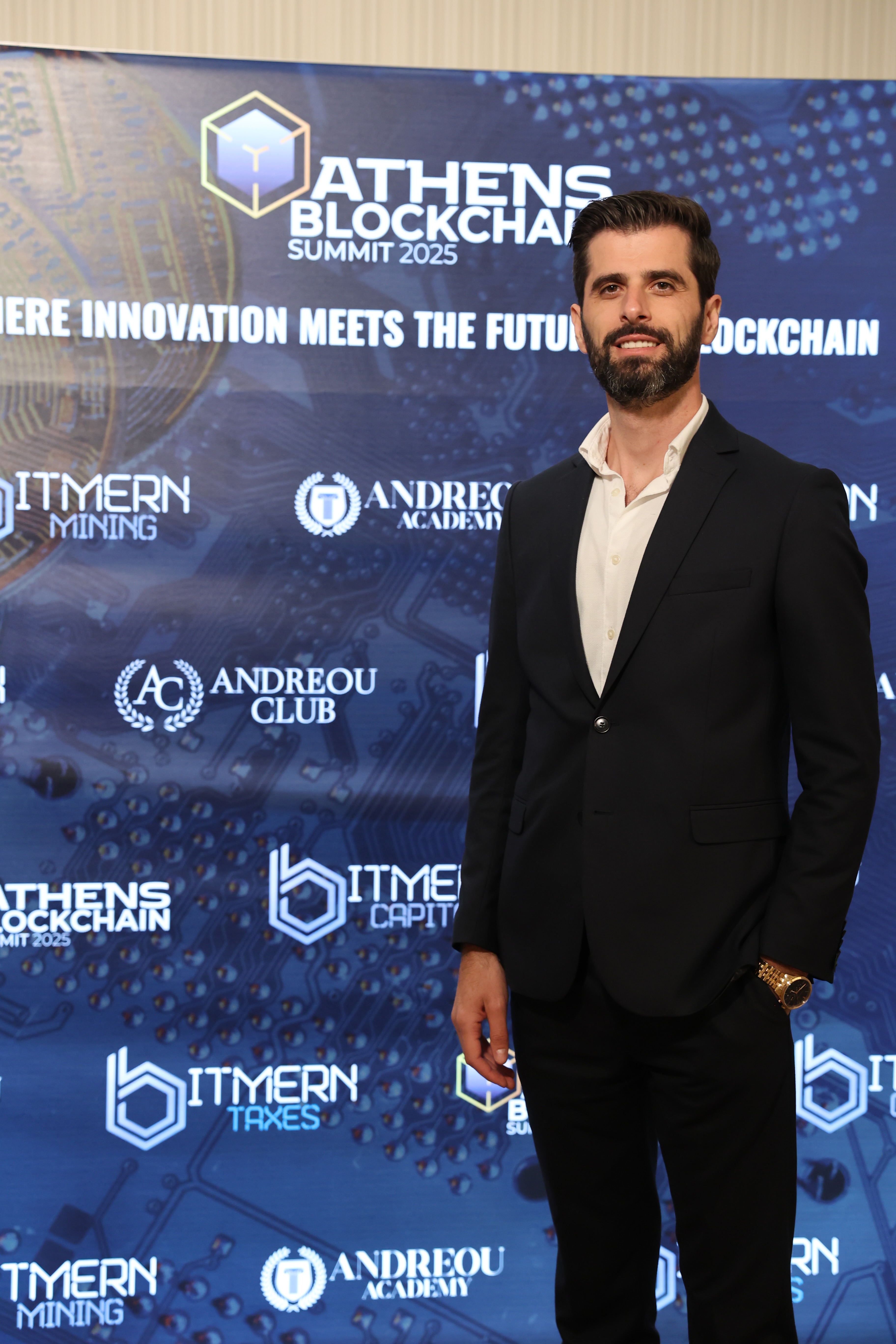 Man in black suit and white shirt standing in front of a screen displaying Athens Blockchain Summit 2025 and sponsor logos.