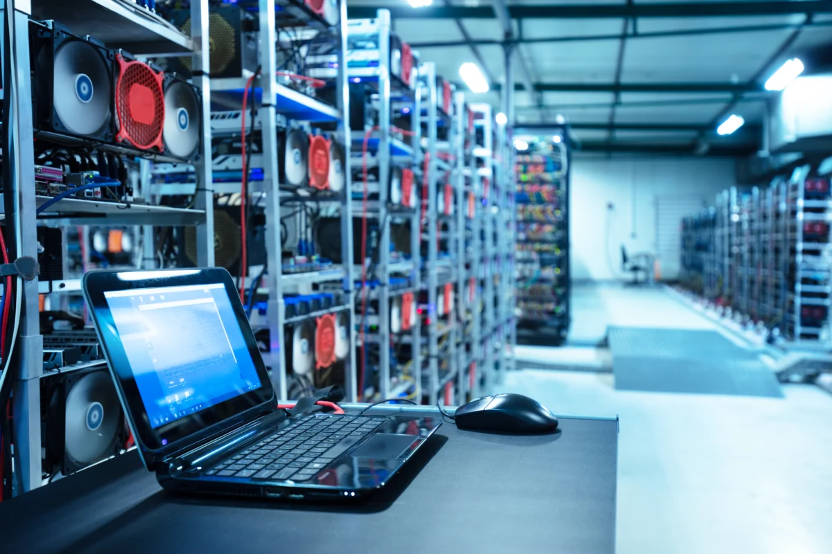 Laptop and mouse on a table in a large cryptocurrency mining farm with rows of GPU rigs and cooling fans.