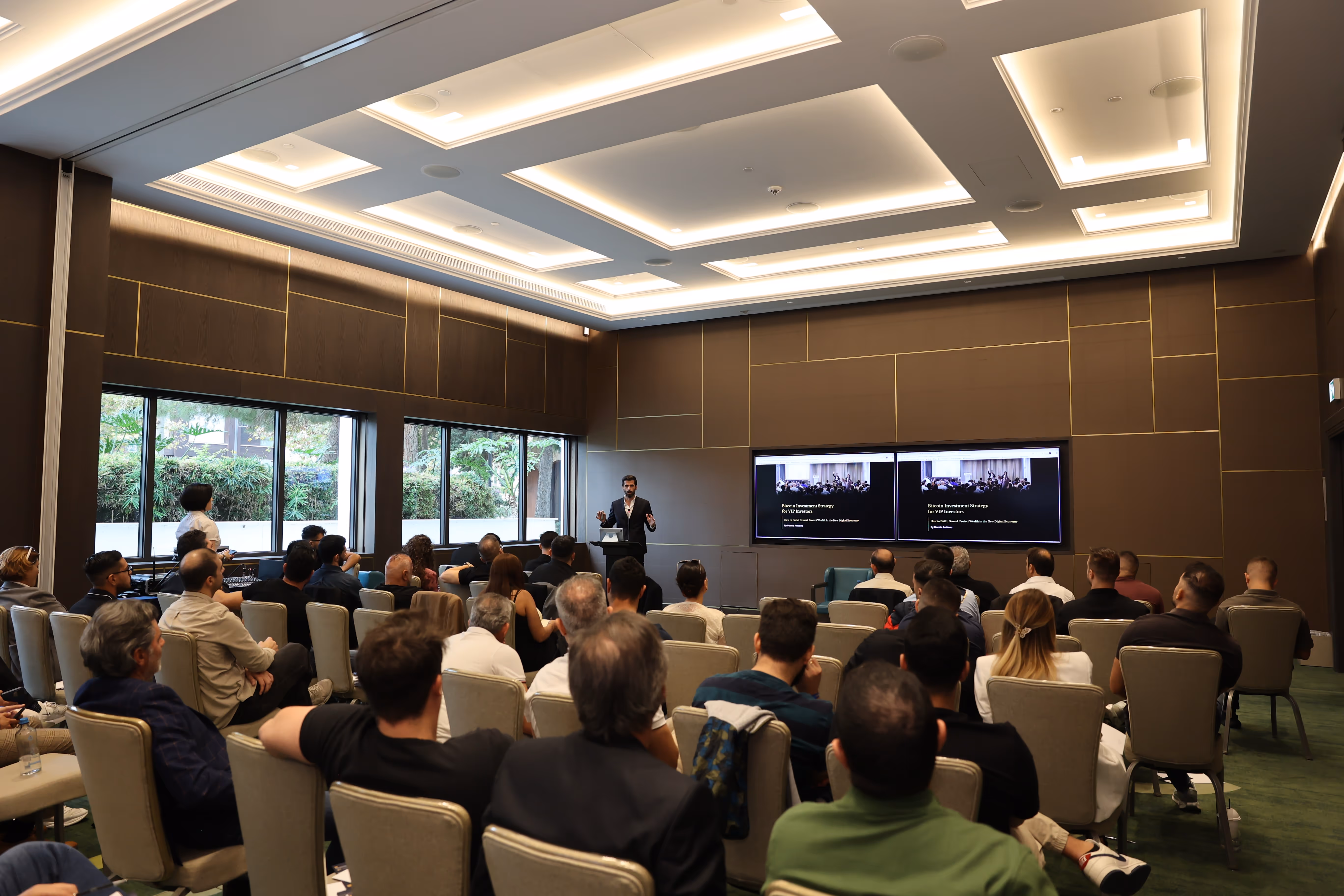 Speaker presenting Bitcoin investment strategy to a seated audience in a modern conference room with two large screens.